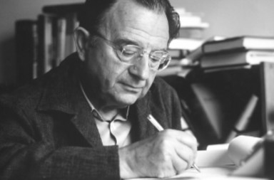 A historical black and white photograph of the psychologist and philosopher Erich Fromm wearing glasses, focused on writing with a pen on a paper at his desk surrounded by books.