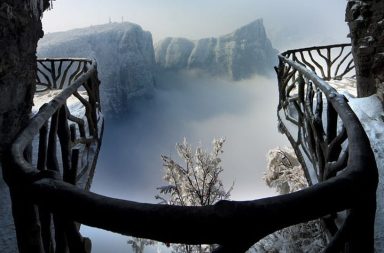 Vista panorâmica a partir de uma passarela com corrimão de madeira na Montanha Tianmen, na China, rodeada por picos cobertos de neve e névoa densa.