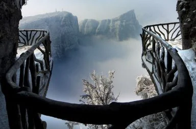 Vista panorâmica a partir de uma passarela com corrimão de madeira na Montanha Tianmen, na China, rodeada por picos cobertos de neve e névoa densa.
