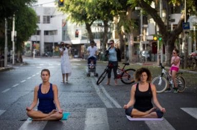 Duas mulheres sentadas na posição de lótus praticam meditação no meio de uma rua deserta, rodeadas por peões com bicicletas ao fundo.