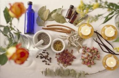 Vários frascos de vidro transparente com essências aromáticas alinhados num balcão de madeira, com folhas verdes e pequenas flores brancas à volta.