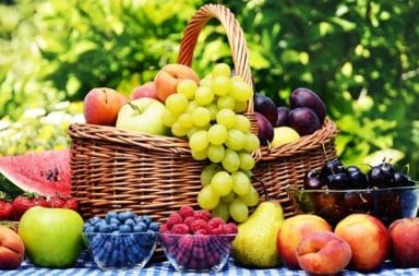 Uma cesta de vime cheia de uvas verdes, pêssegos e maçãs, cercada por tigelas com mirtilos, framboesas e cerejas sobre uma mesa