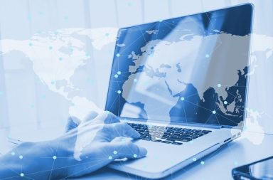 Uma mão humana operando o teclado de um computador portátil aberto com uma sobreposição gráfica azul em transparência que exibe um mapa-múndi interconectado por pontos e linhas de rede.