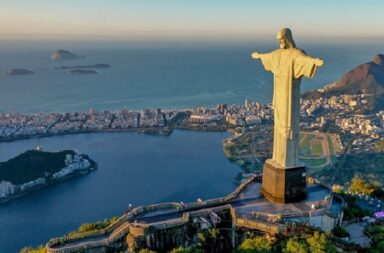 Vista aérea da estátua do Cristo Redentor de braços abertos no topo do Corcovado, com a cidade do Rio de Janeiro e o oceano ao fundo.