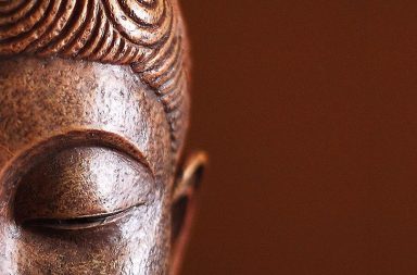 Perfil em close-up de uma estátua de Buda em tons de bronze e madeira, com os olhos fechados num estado de meditação profunda contra um fundo castanho liso.