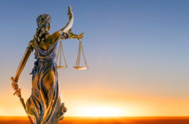 Estátua de bronze de Têmis, a Deusa da Justiça, vista de costas, segurando uma espada na mão direita e uma balança de dois pratos erguida na mão esquerda, posicionada contra um céu ao amanhecer com tons de azul e um horizonte dourado iluminado pelo sol.