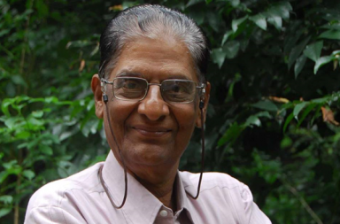 A portrait photograph of the theosophist N. C. Ramanujachary smiling, wearing eyeglasses with a neck strap and a light pink shirt, framed by a dark green borders overlay with a natural green foliage backdrop.