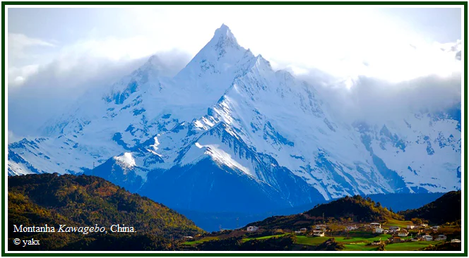 Pico nevado e imponente da montanha Kawagebo, sob nuvens e acima de colinas verdes com pequenas casas, ilustrando o degelo no Tibete.