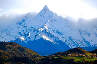 Pico nevado e imponente da montanha Kawagebo, sob nuvens e acima de colinas verdes com pequenas casas, ilustrando o degelo no Tibete.