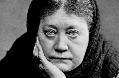 Retrato fotográfico histórico em plano aproximado e a preto e branco de Helena Blavatsky, coberta com um xaile escuro sobre a cabeça, apoiando o rosto pensativo na mão direita com um olhar fixo e penetrante direcionado para a câmara.
