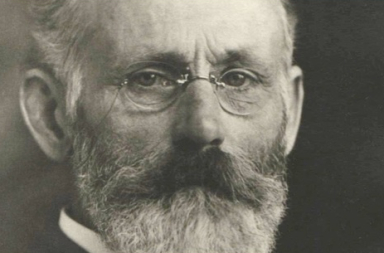 Retrato fotográfico histórico em plano aproximado e a preto e branco do teosofista Robert Crosbie, exibindo os seus óculos de lentes ovais, cabelo curto e uma barba e bigode volumosos e grisalhos, cujo ensinamento analisa o dogmatismo religioso.