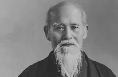 Retrato fotográfico histórico de Morihei Ueshiba, o fundador cujos ensinamentos estabeleceram a filosofia do aikidô, exibindo o seu rosto sereno com uma longa barba e bigode brancos, contra um fundo neutro de estúdio.