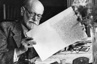 Retrato fotográfico histórico a preto e branco de Sigmund Freud, o fundador da psicanálise, sentado à sua secretária com óculos de leitura, concentrado a analisar um manuscrito em papel que segura com a mão esquerda.