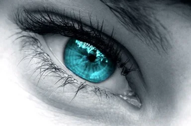 Fotografia artística macro e em plano aproximado de um olho humano com uma íris azul-turquesa brilhante e cílios longos e pretos em destaque, editada em tons suaves de cinzento e enquadrada por uma fina moldura verde, ilustrando a perceção da beleza.