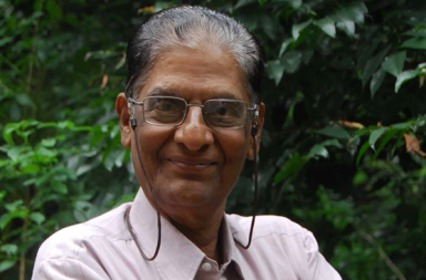 Retrato fotográfico em plano médio do teosofista N. C. Ramanujachary sorridente, usando óculos com cordão e camisa clara, posando contra um fundo denso de vegetação e folhas verdes escuras, cujo artigo analisa os erros em relação aos Mestres de sabedoria.