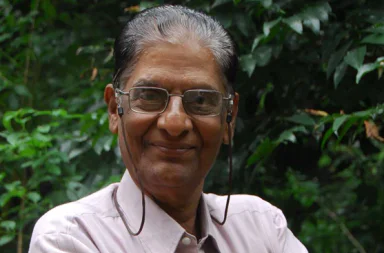 Retrato fotográfico em plano médio do teosofista N. C. Ramanujachary sorridente, usando óculos com cordão e camisa clara, posando contra um fundo denso de vegetação e folhas verdes escuras, cujo artigo analisa os erros em relação aos Mestres de sabedoria.
