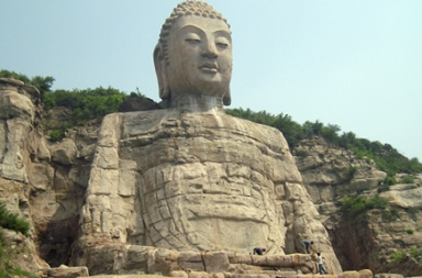 Vista frontal da colossal estátua de pedra do Buda Gigante de Mengshan, esculpida diretamente na encosta de uma montanha rochosa, exibindo uma expressão serena e meditativa com olhos semicerrados, rodeada por vegetação esparsa nas falésias.