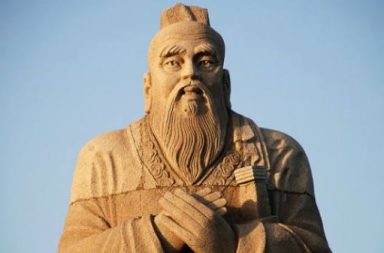 Plano médio da colossal estátua de pedra de Confúcio com longas barbas e mãos cruzadas sobre o peito, cuja sabedoria tradicional está preservada nos analectos, isolada contra um fundo de céu azul limpo.