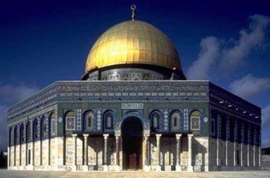 Vista frontal da Cúpula da Rocha em Jerusalém, exibindo a sua imensa cúpula dourada brilhante e a fachada octogonal detalhada com azulejos azuis e colunas clássicas sob um céu limpo, ilustrando o artigo sobre o islamismo.
