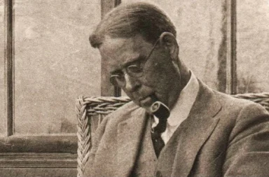 Fotografia histórica em tom sépia do teosofista John Garrigues de perfil, usando óculos e casaco, sentado numa cadeira de vime enquanto fuma cachimbo.