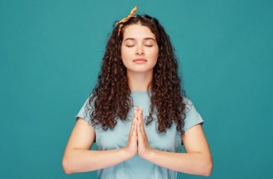 Mulher de olhos fechados fazendo o gesto de namastê com as mãos unidas em meditação, com fundo azul.