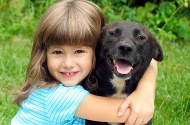 Uma menina sorridente abraça um cão preto num relvado verde, ilustrando o laço puro e a força da confiança.