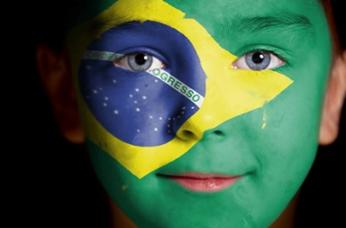 Plano aproximado do rosto de uma criança com os olhos azuis e a bandeira do Brasil pintada na face, simbolizando o povo brasileiro.