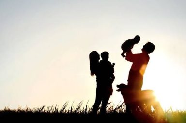 Silhueta escura ao pôr do sol de uma família num campo de relva, mostrando o pai a erguer um bebé no ar, a mãe a segurar uma criança ao colo e o cão de estimação ao lado, envoltos por um forte brilho solar contra o céu claro.