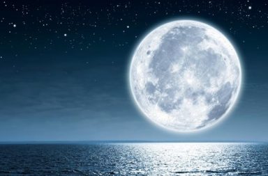 Fotografia da lua cheia em plano aproximado com luz prateada a refletir no mar e horizonte noturno.