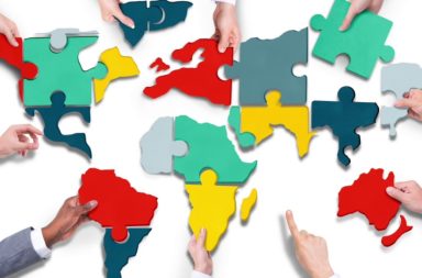 Várias mãos de diferentes tons de pele encaixando peças de puzzle coloridas que formam o mapa-múndi sobre um fundo branco.