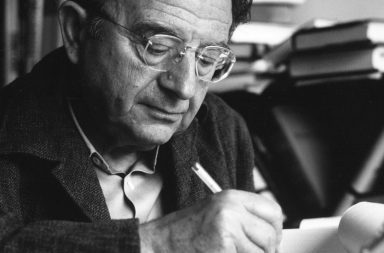 Retrato fotográfico histórico a preto e branco do psicanalista Erich Fromm, usando óculos e casaco escuro, concentrado a escrever com uma caneta num papel sobre a secretária, cuja análise psicológica ajuda a compreender o papel do ritual religioso na sociedade humana.